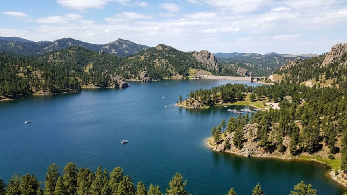 Blue lake surrounded by forested hills and rocky cliffs, with a distant dam at the far end.