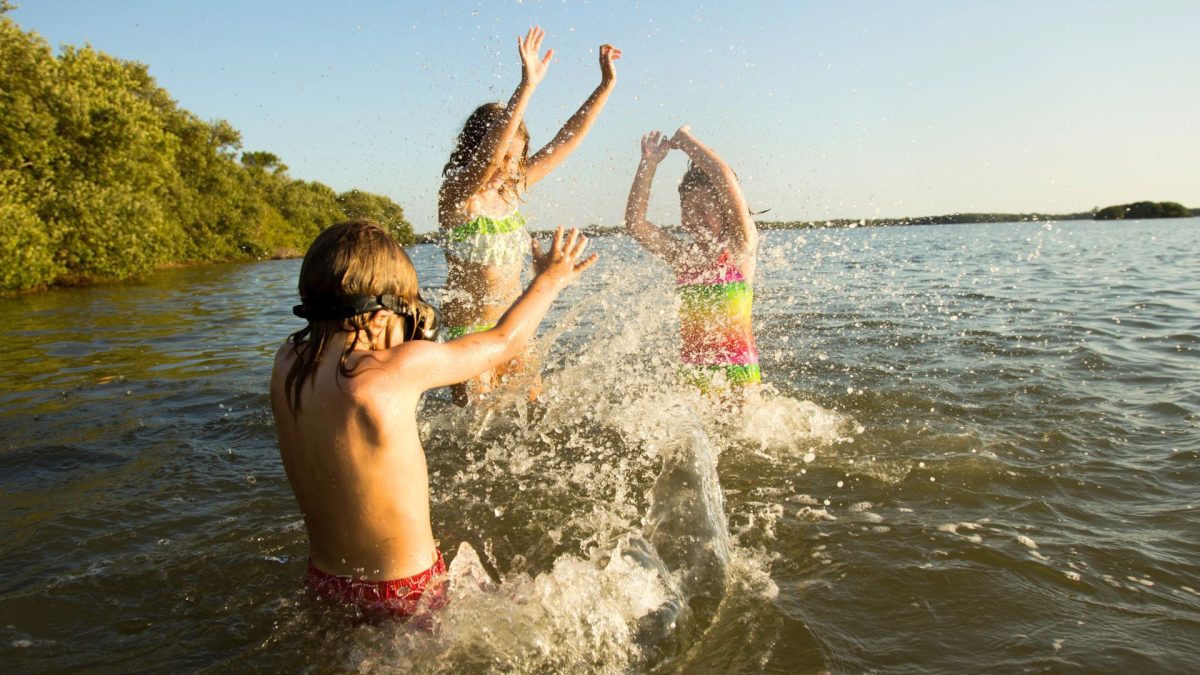 Children in swimsuits splash and jump in a lake while water sprays around them on a sunny day, with trees in the background.