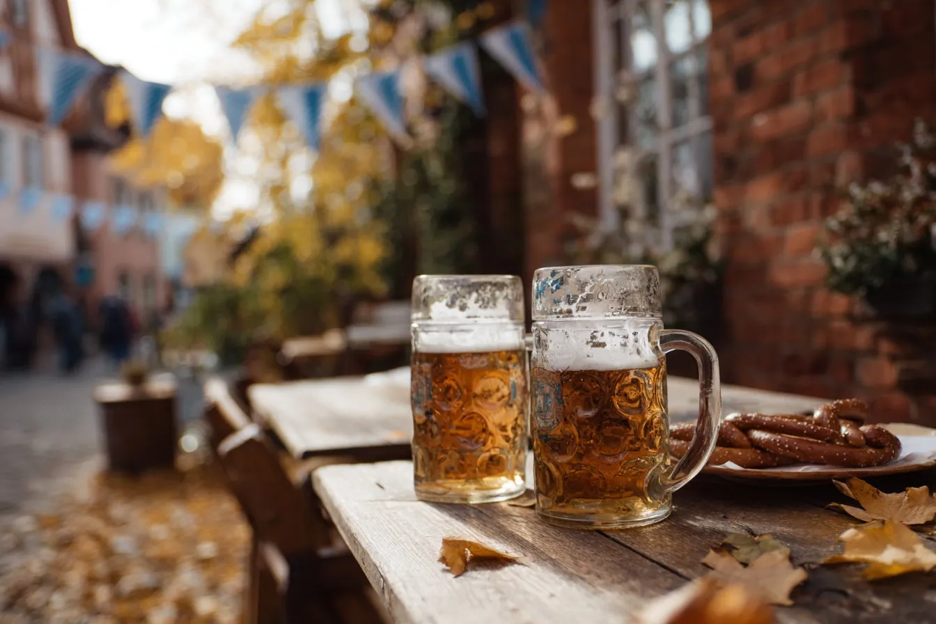 Two beer steins and soft pretzels on a wooden table outdoors with autumn leaves and blue and white bunting