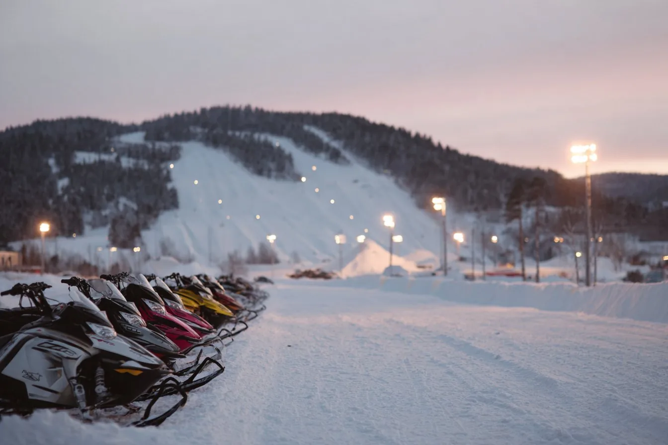 Row of race snowmobiles parked on snow beside a lit snocross course at dusk