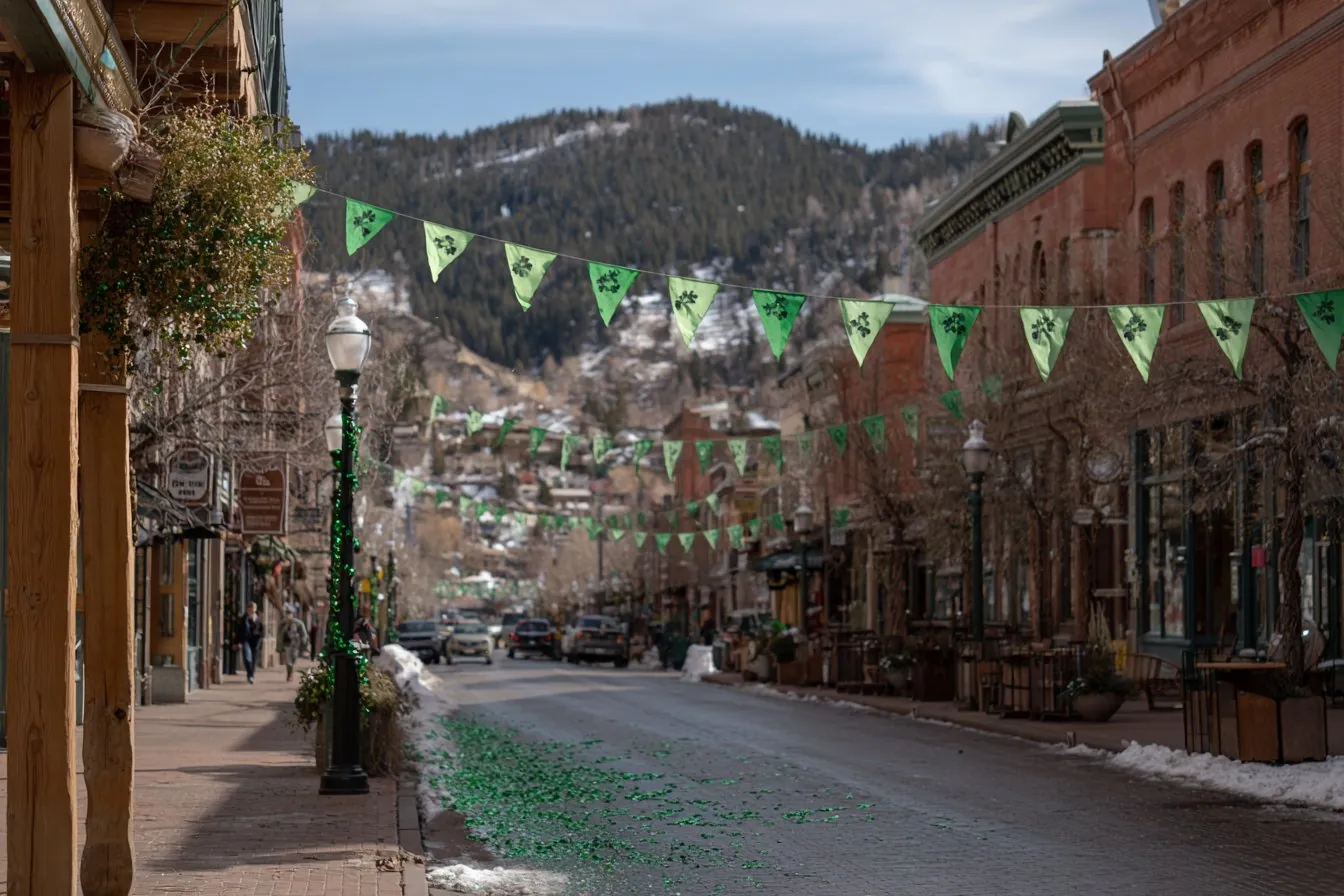Green shamrock pennant banners strung over a historic brick main street with green confetti on the road and snowy pine hills behind